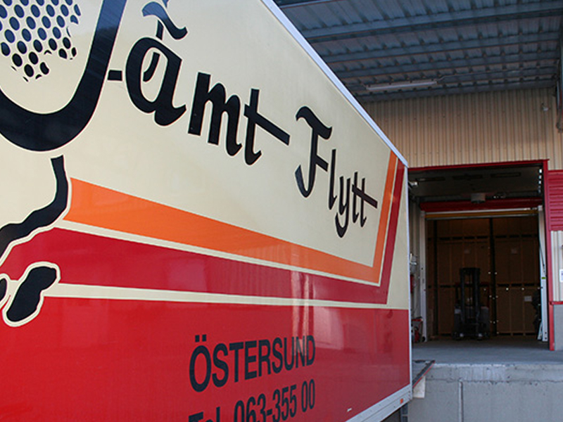 jamtflytt-lokaler2 Närbild på Jämt Flytt-lastbil vid lastkaj, med öppen port till lagerutrymme där träboxar och truck syns i bakgrunden.