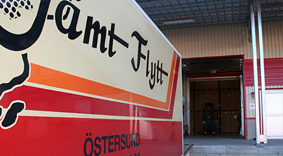 Magasinering_580 Närbild på Jämt Flytt-lastbil vid lastkaj, med öppen port till lagerutrymme där träboxar och truck syns i bakgrunden.